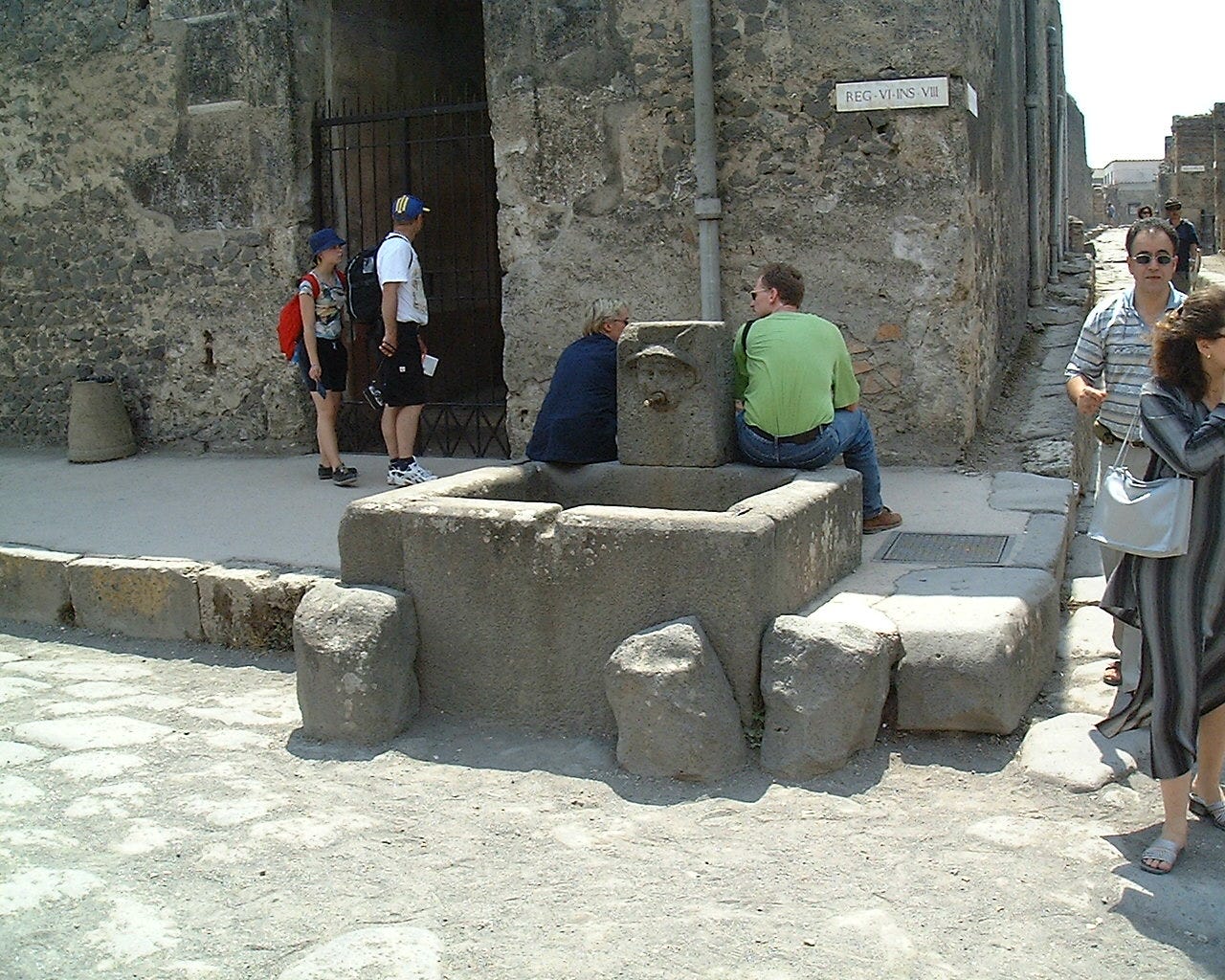 Tourists gather on a street corner in Pompeii. A couple perch on the stone basin of an ancient fountain. A head of Mercury, wearing his winged hat forms the spout with the water coming out of his mouth.