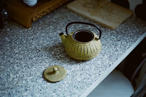 1) A sage green tea pot sitting next to it's lid on a black & white speckled counter. 2) Two dark purple and white china tea cups filled with tea & orange slices, sitting next to a sprig of holly berries. 3) A frog in a red & white mushroom outfit, resting atop a beige-coloured pumpkin. 4) Out a window, a golden sunrise across a city-scape. 5) A stack of six artistically decorated soda cans. 6) 5 old & differently shaped keys.