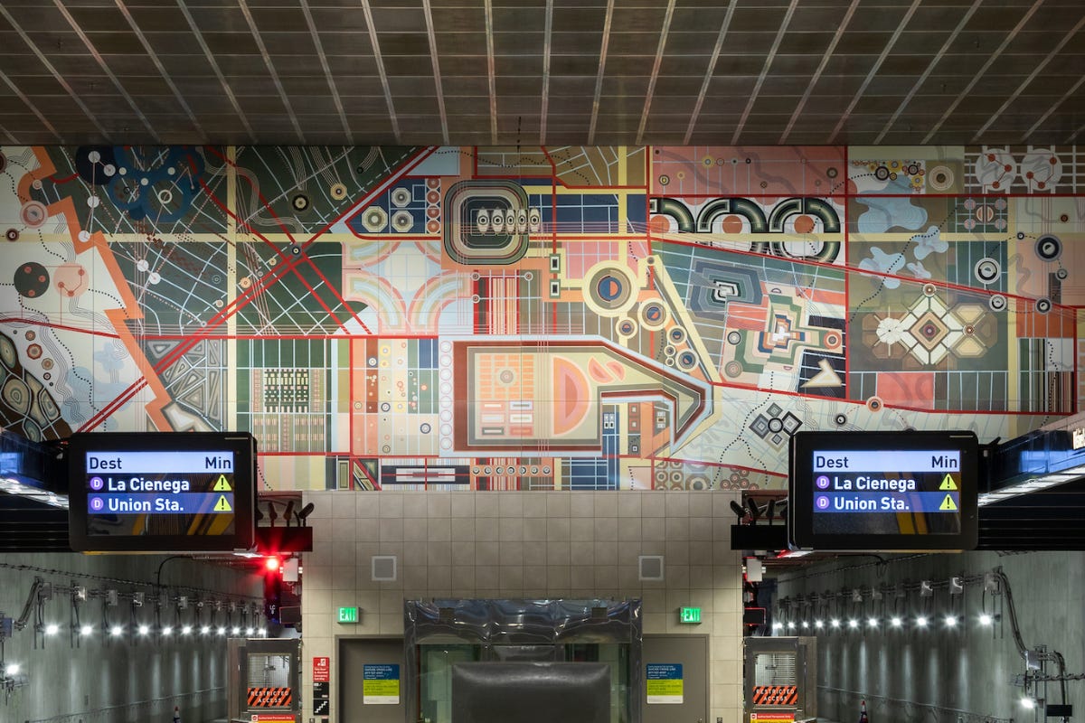 A colorful horizontal station artwork stretches above train signs and platform lights, combining abstract maps, architectural forms, patterns, and layered city imagery. A colorful horizontal station artwork stretches above train signs and platform lights, combining abstract maps, architectural forms, patterns, and layered city imagery.