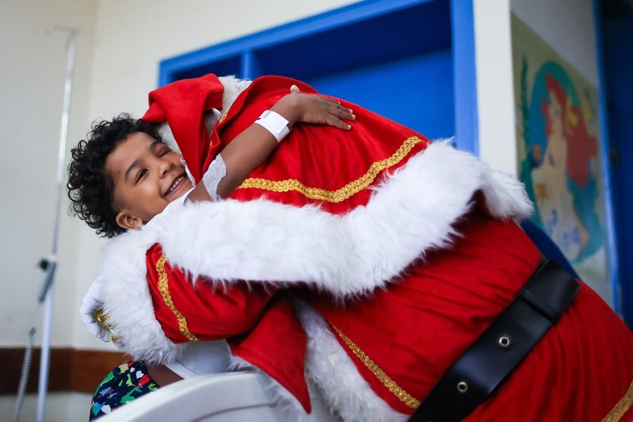 Dressed as Santa Claus, a person leans over to give a big hug to a young patient in a hospital bed who smiles and hugs back. Dressed as Santa Claus, a person leans over to give a big hug to a young patient in a hospital bed who smiles and hugs back.