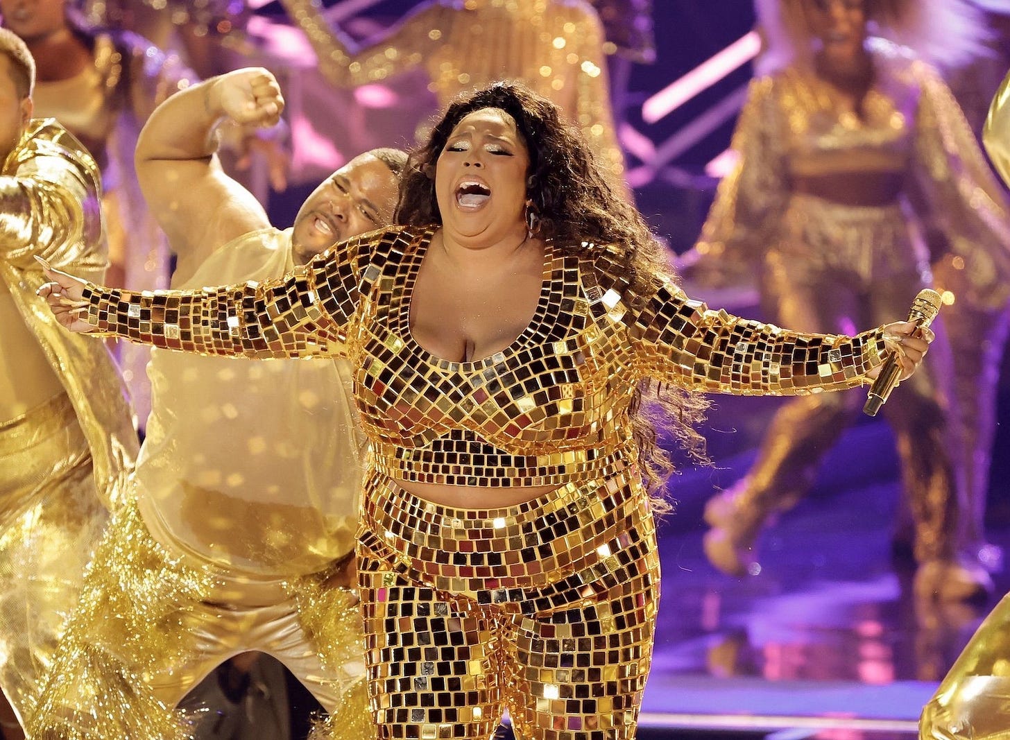 Lizzo performing onstage in a gold mirrorball bodysuit, singing powerfully under bright lights with dancers behind her. A mid-distance shot emphasizing her presence and performance rather than her body.