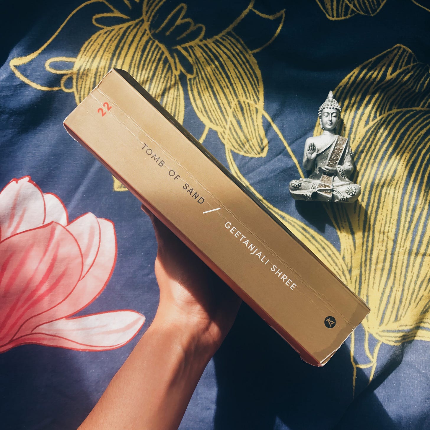 holding “Tomb of Sand” diagonally to show the spine of the book alongside a small Buddha statue. Navy blue and yellow background holding “Tomb of Sand” diagonally to show the spine of the book alongside a small Buddha statue. Navy blue and yellow background
