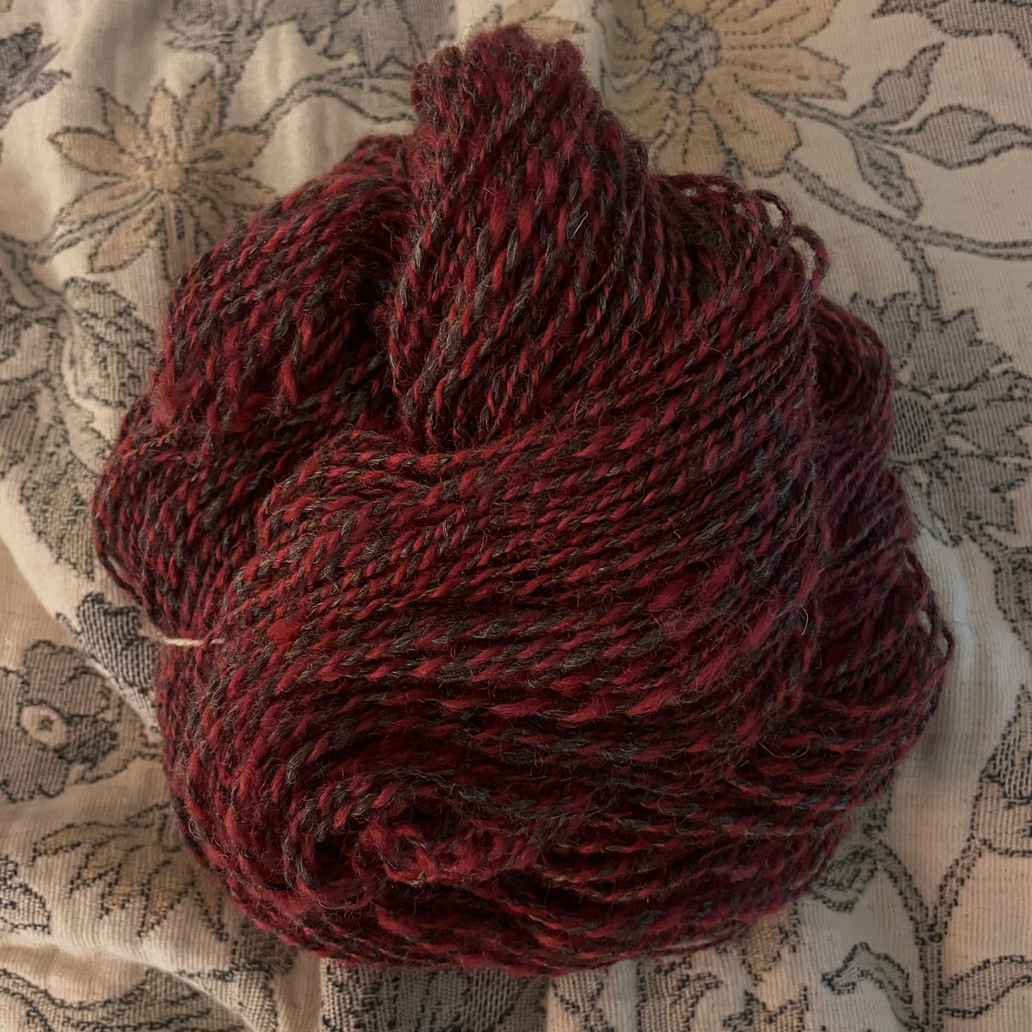 An image of a pile of handspun yarn. It is dark grey and red marled yarn that sits on a floral blanket.