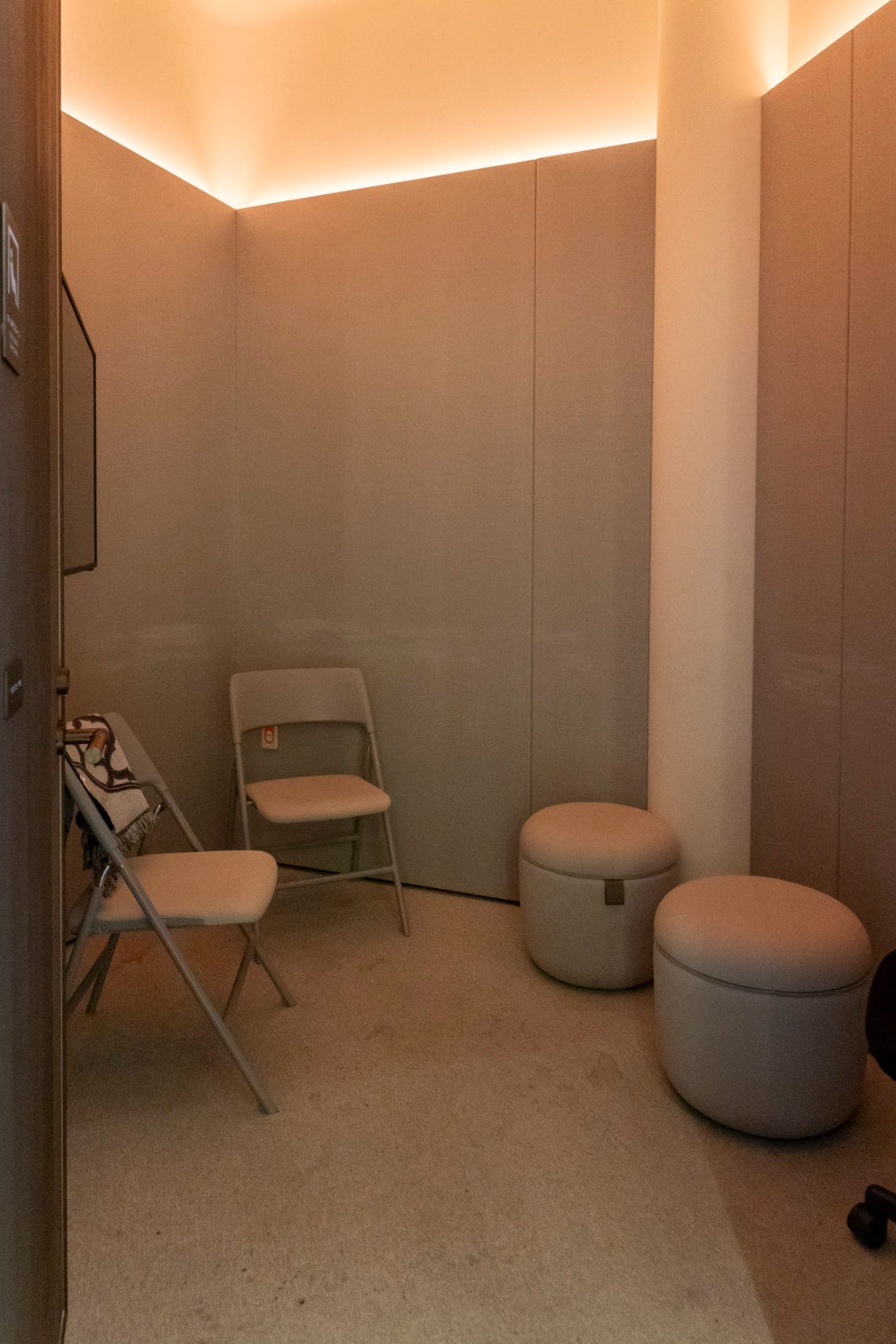 Quiet room with soft lighting, two folding chairs, and two round cushioned stools against beige walls.