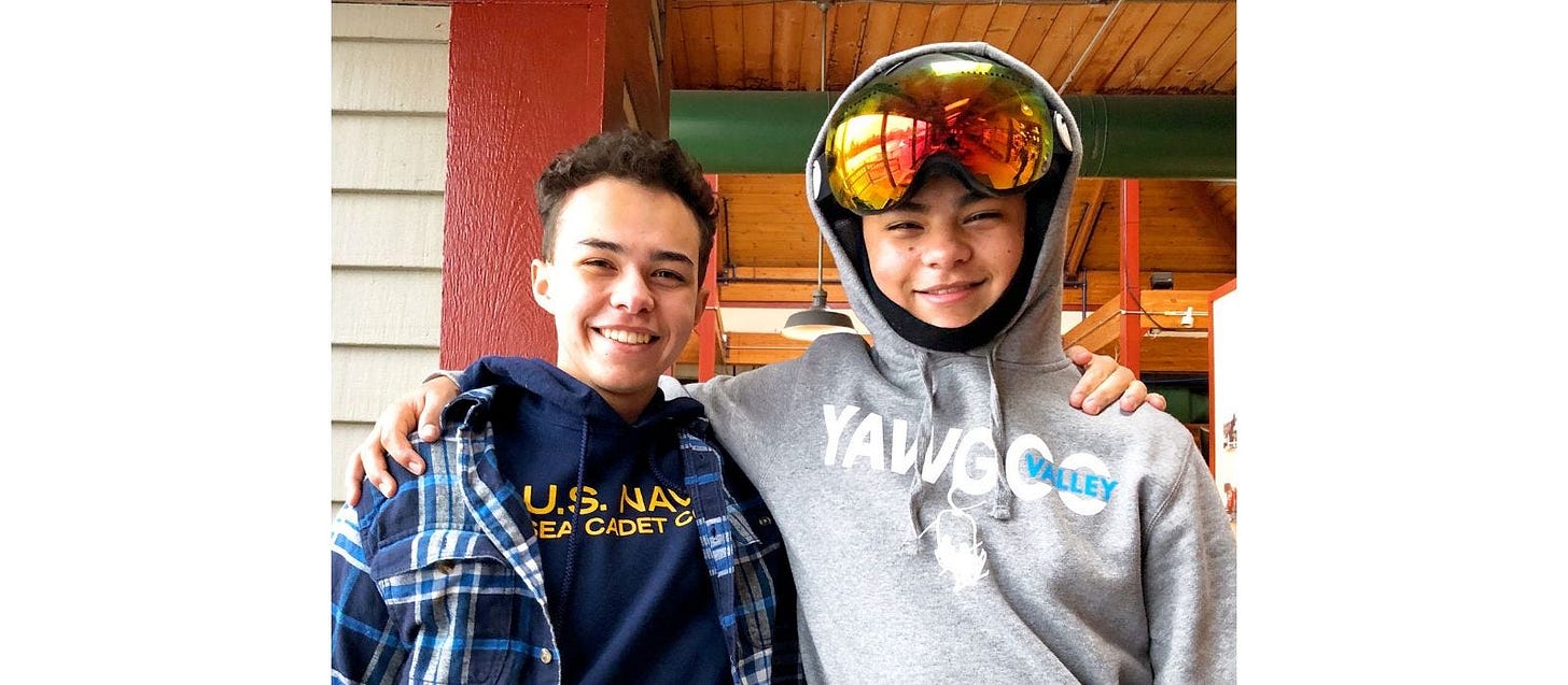 2 high school boys with arms around each other’s shoulders, both wear warm sweatshirts, one has on a ski helmet and goggles on top of his head
