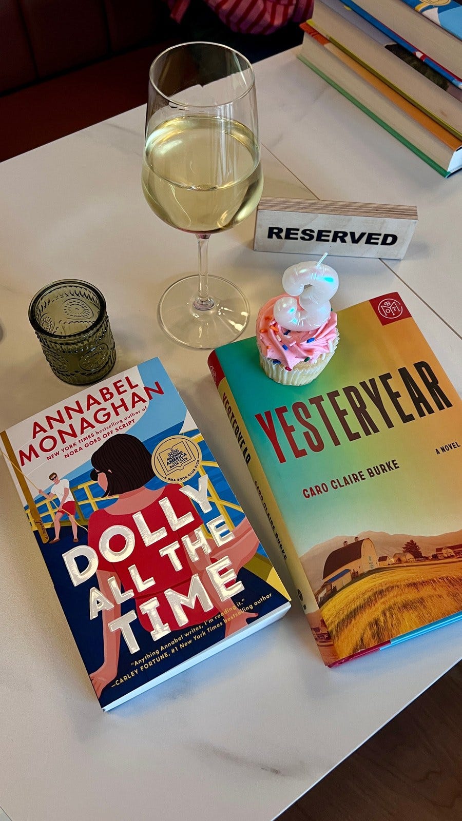 A book club table with a glass of white wine, a RESERVED sign, a cupcake, and two novels.