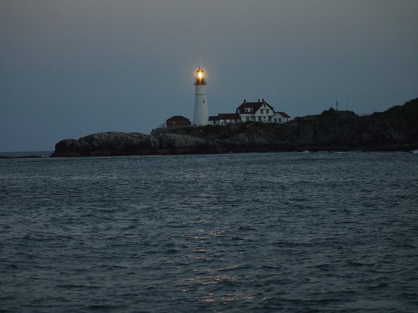 Dim photo of a lighthouse and some small houselike buildings on a rocky coast. The lighthouse beam is aimed directly at the camera. The sea and sky are gray-blue. The rocks are black.