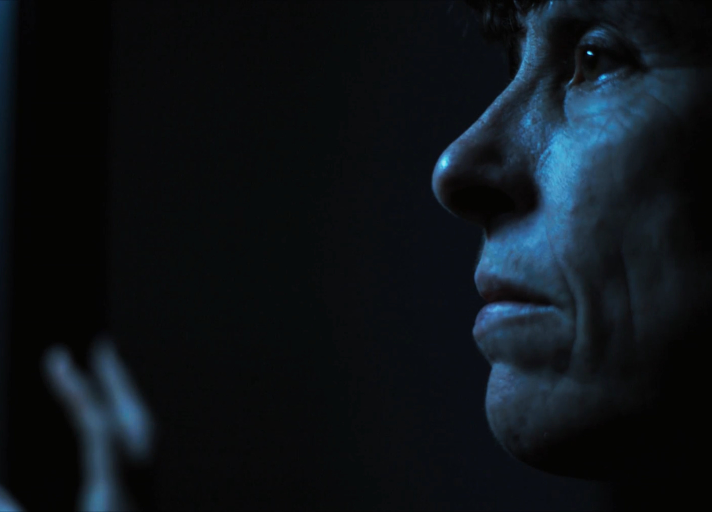 Andrea Gibson in a dark room staring out the window with blue moonlight drenching their sad face