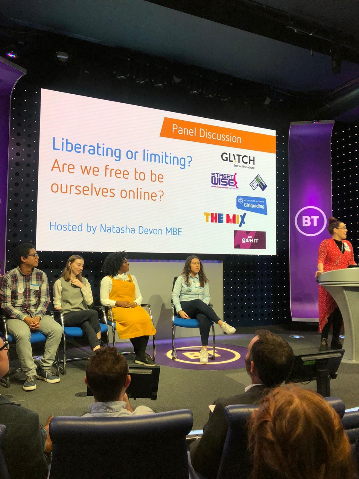 The image shows a panel discussion titled "Liberating or Limiting? Are we free to be ourselves online?" with several speakers sitting on stage. The panel is hosted by Natasha Devon MBE, and the logos of various organizations such as GLITCH, StreetWise, The Mix, and Girlguiding are displayed. The panelists include a diverse group of individuals, one of whom appears to be Seyi Akiwowo, wearing an orange dress. The discussion is taking place at a BT event.