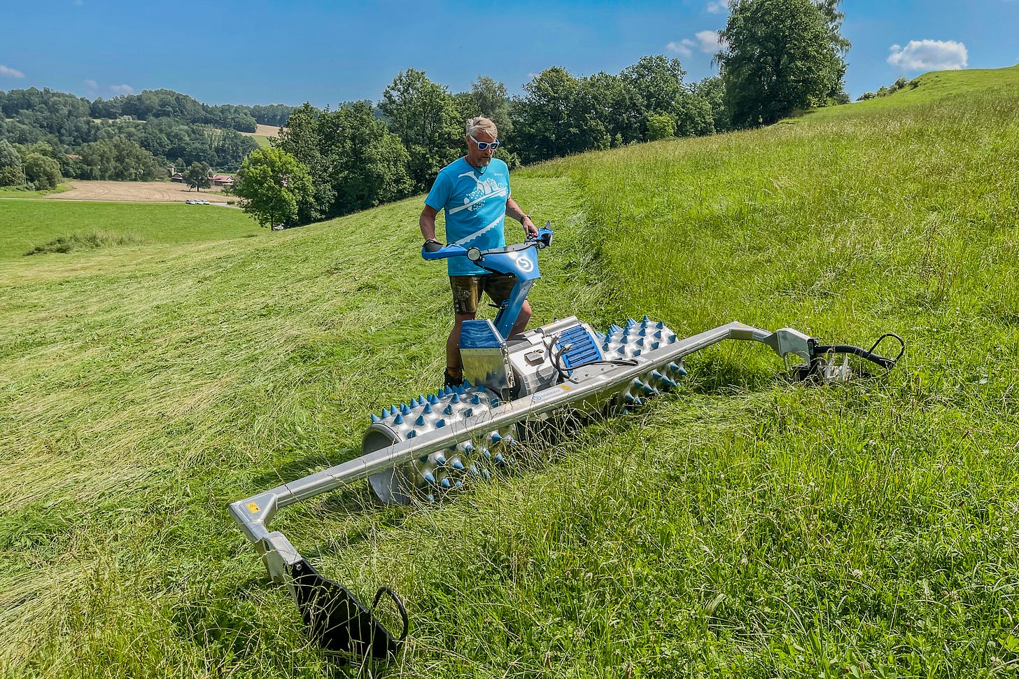 Ein Mann in blauem T-Shirt und Sonnenbrille arbeitet mit einem einachsigen Brielmaier-Geräteträger an einer Hangwiese. Vor ihm mäht die Maschine das Gras mit seitlich ausladendem Balken; hinter dem Gerät ist die Fläche bereits sauber geschnitten. Im Hintergrund liegen Bäume, sanfte Hügel und einzelne Höfe unter blauem Himmel. Ein Mann in blauem T-Shirt und Sonnenbrille arbeitet mit einem einachsigen Brielmaier-Geräteträger an einer Hangwiese. Vor ihm mäht die Maschine das Gras mit seitlich ausladendem Balken; hinter dem Gerät ist die Fläche bereits sauber geschnitten. Im Hintergrund liegen Bäume, sanfte Hügel und einzelne Höfe unter blauem Himmel.