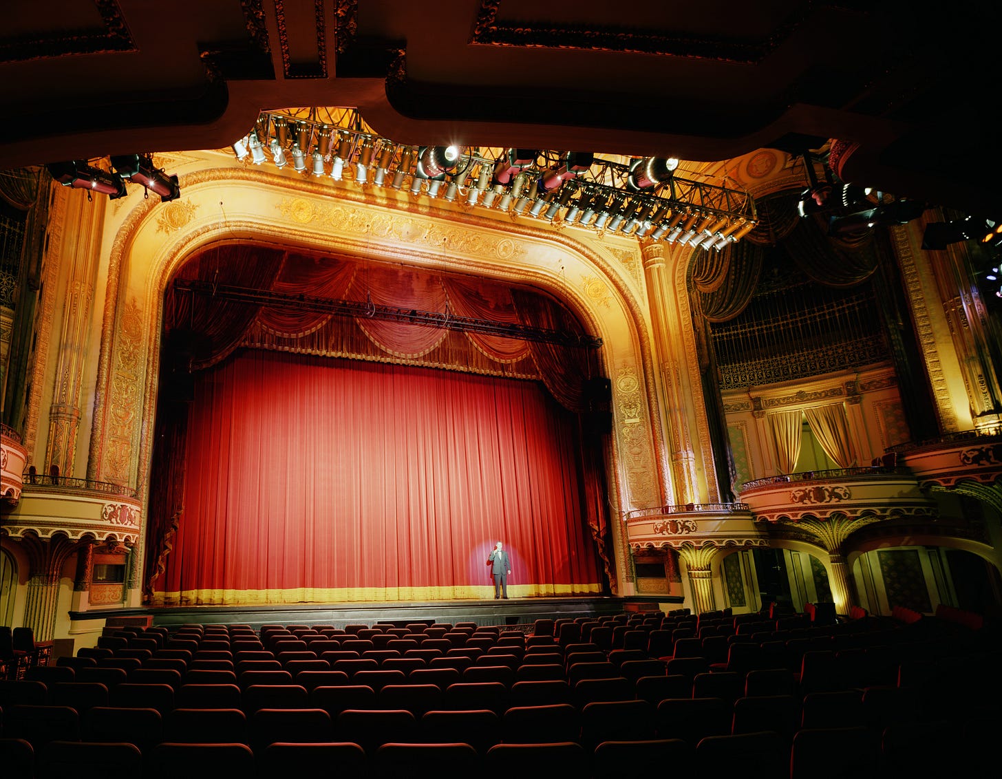 Classic  early 20th century performance theatre with one man on stage in a spotlight. Gold walls, red curtain, balconies and loges