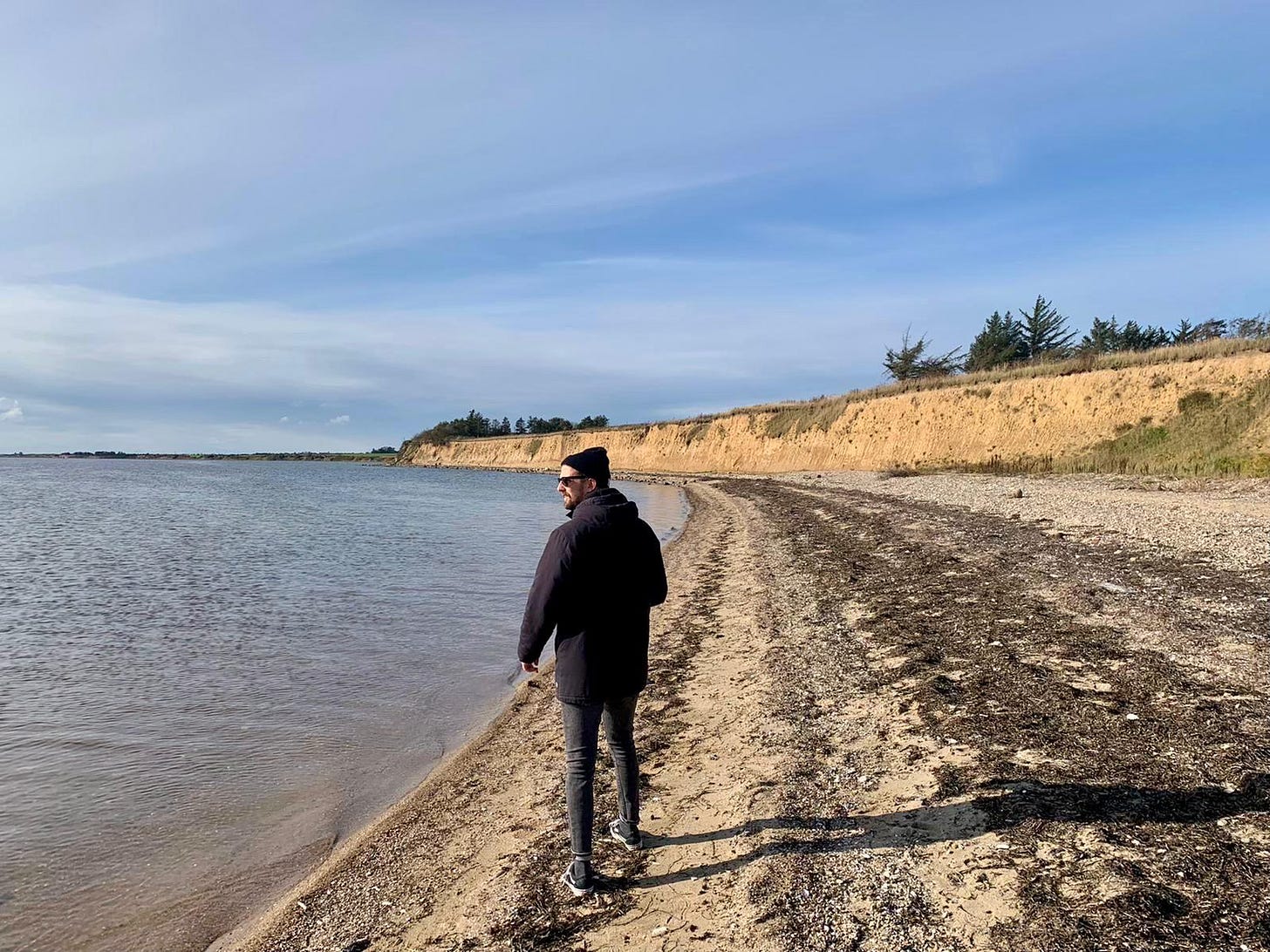 A person walking alone on a cold beach in Denmark wearing a coat, the grey sea and overcast sky stretching behind them. A person walking alone on a cold beach in Denmark wearing a coat, the grey sea and overcast sky stretching behind them.