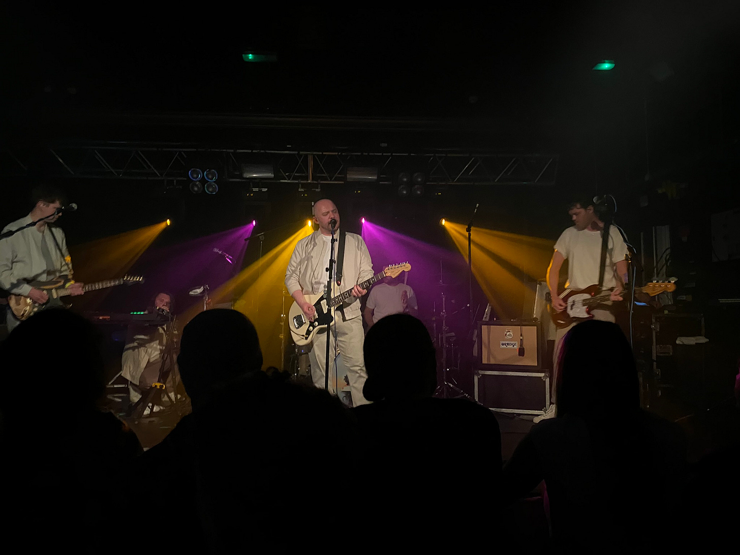 Jonathan Higgs, the lead singer of Everything Everything, is playing guitar with the rest of the band. He's being backed up by orange and purple lighting. Some of the crowd in front of Avery is visible in silhouette. Jonathan Higgs, the lead singer of Everything Everything, is playing guitar with the rest of the band. He's being backed up by orange and purple lighting. Some of the crowd in front of Avery is visible in silhouette.