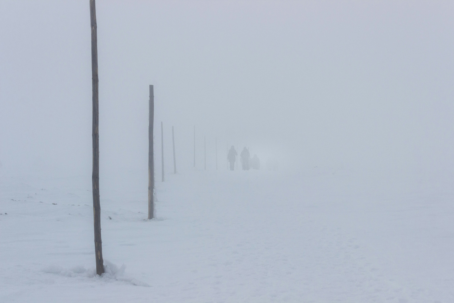 White ground meets white as far as the eye can see in the sky, with a few old utility poles fading in the near distance. Almost not visible are two people walking away from the camera in the snow.