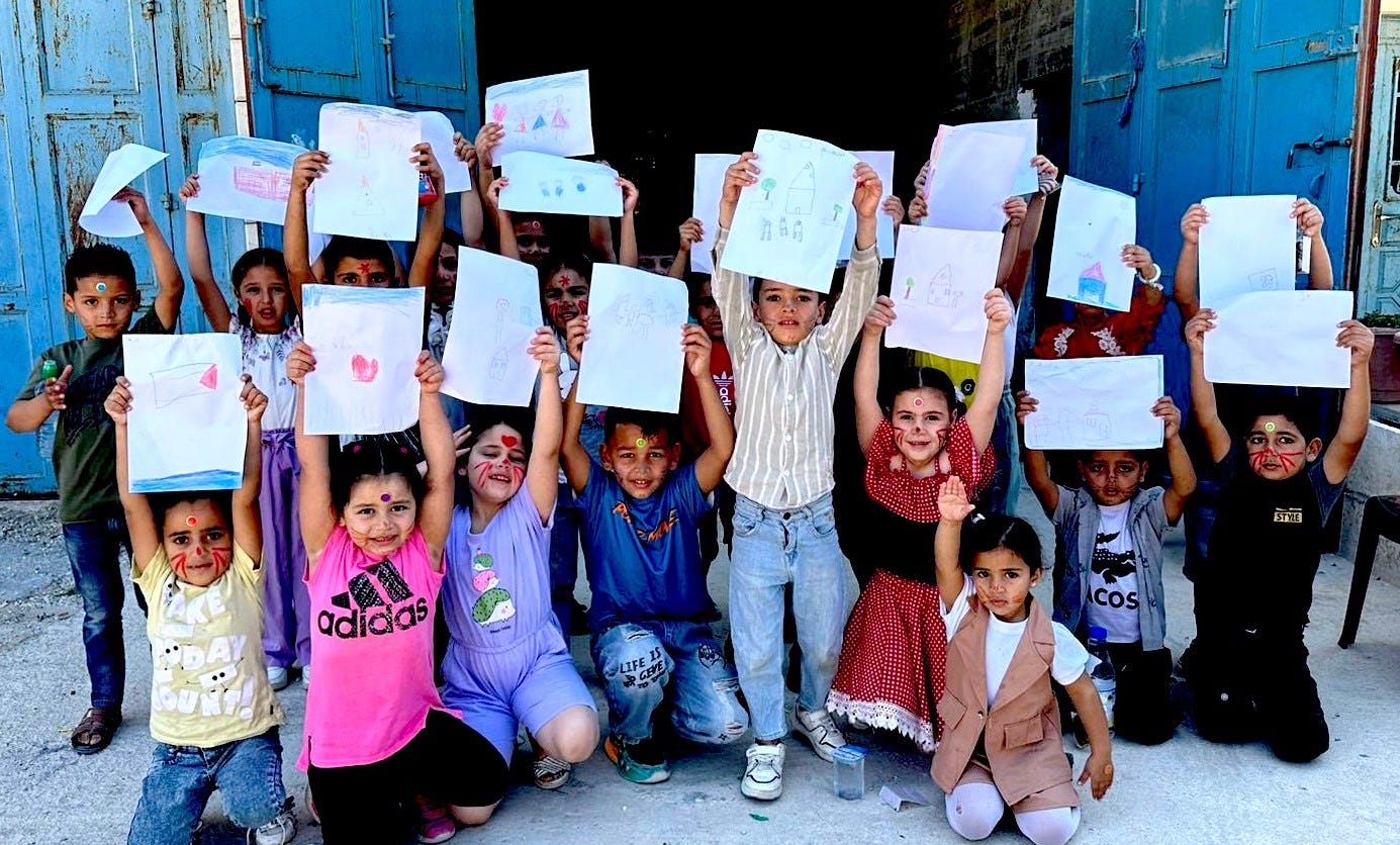 A group of young Palestinian children holding up the drawings they have made, their faces are painted with hearts, cat whiskers, and other playful designs.
