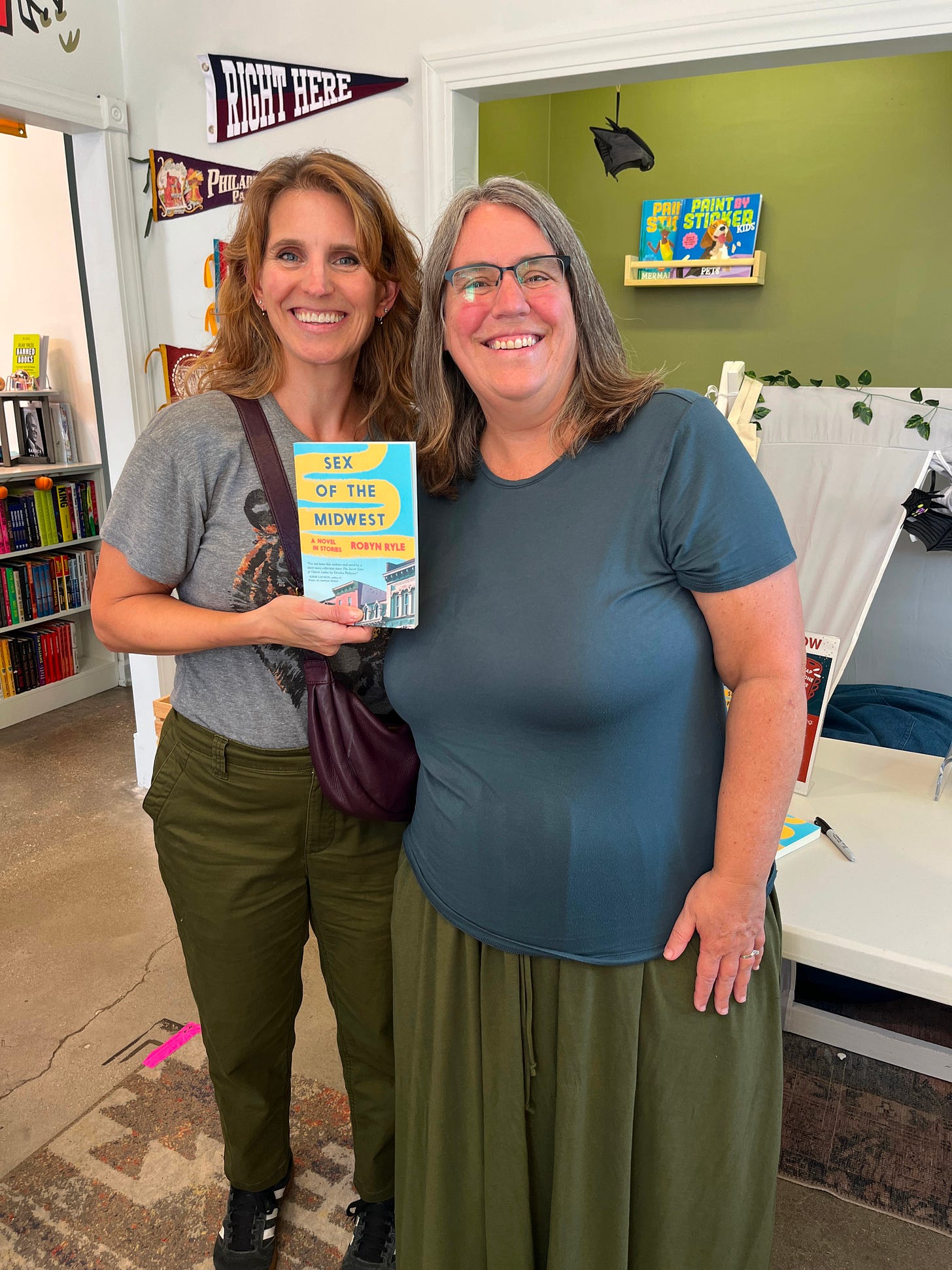 Robyn and Kate standing together, Kate holding up a copy of Sex of the Midwest, both of them smiling