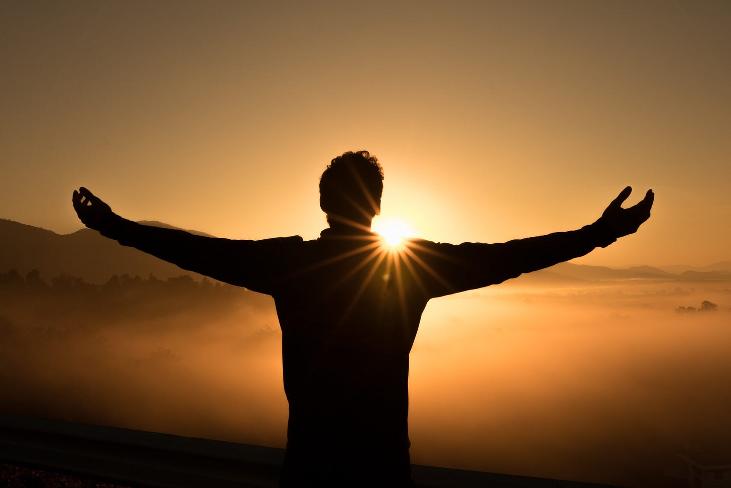 The silhouette of a man with arms outstretched. In front of him is a sunrise and orange sky with hills in the distance.
