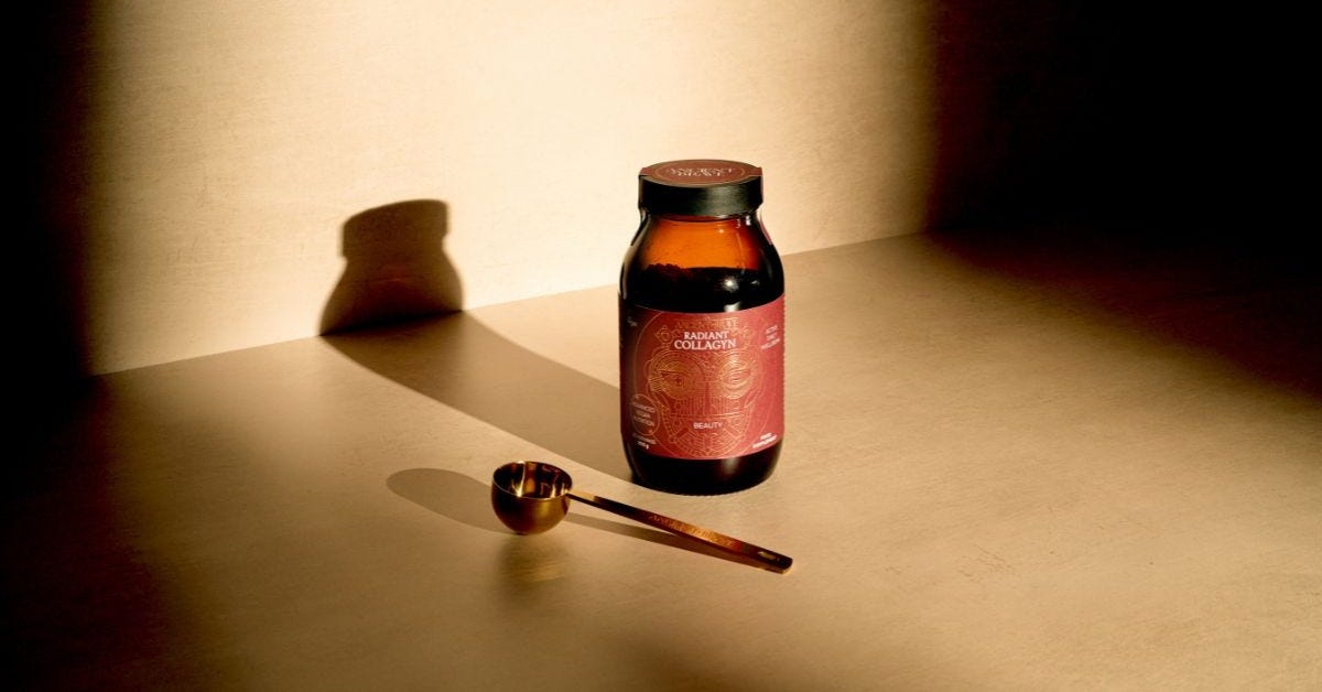 A jar of Radiant Collagyn alongside a golden ritual scoop, in a beam of sunlight on a neutral floor