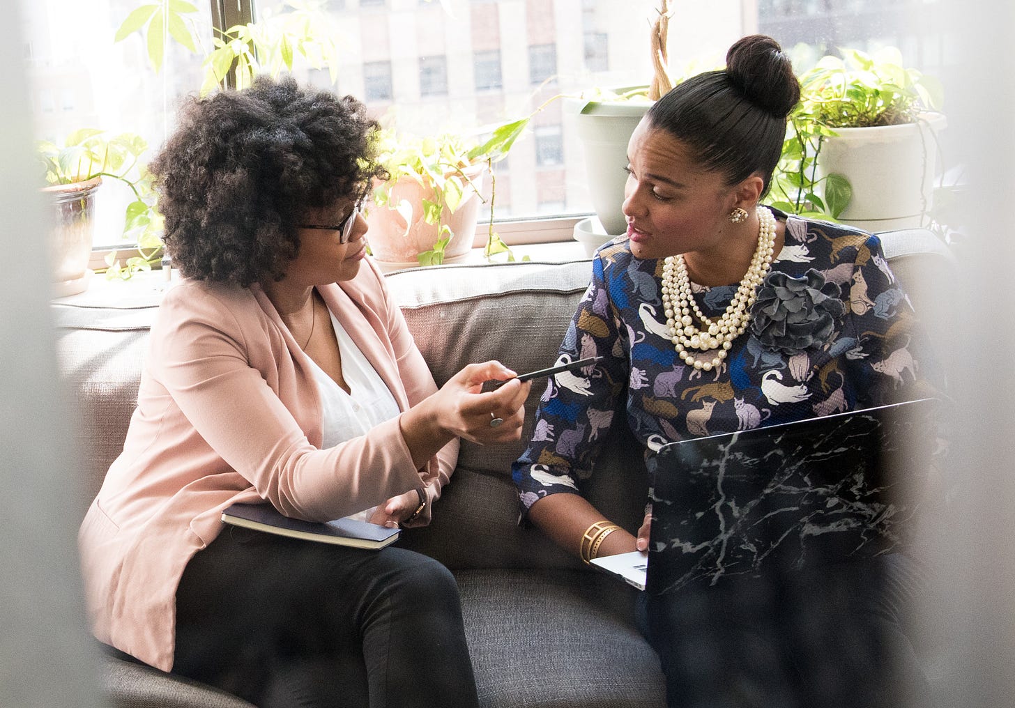 Two women sit next to each other on a couch. The woman on the right seems older, as if a mentor or supervisor. She gestures towards the laptop screen of the woman on the right, as if providing the second woman with advice.