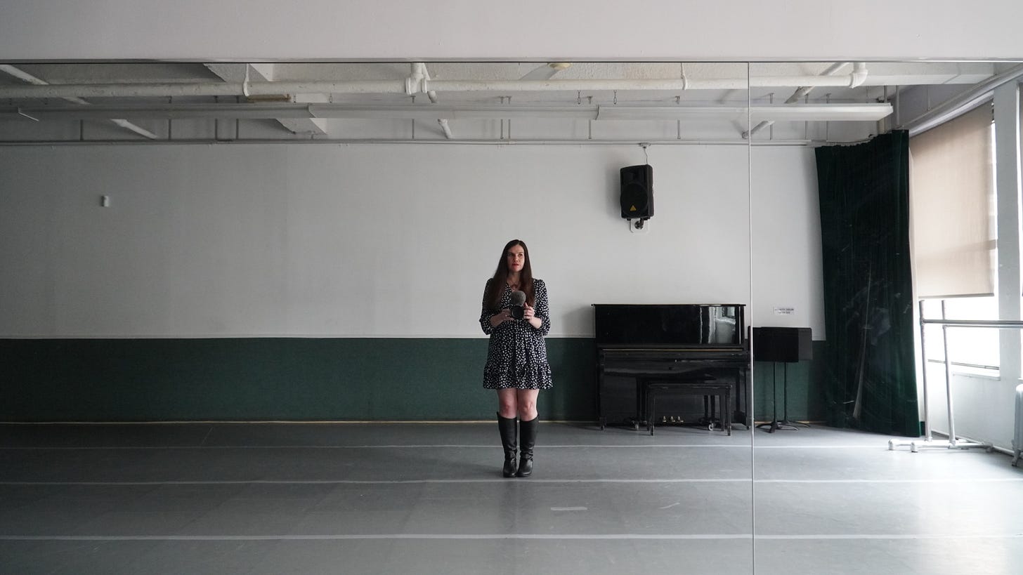 A woman stands in front of a mirror in a rehearsal room. She looks tense and stands to the side. The room is empty and there is a piano behind her. A woman stands in front of a mirror in a rehearsal room. She looks tense and stands to the side. The room is empty and there is a piano behind her.