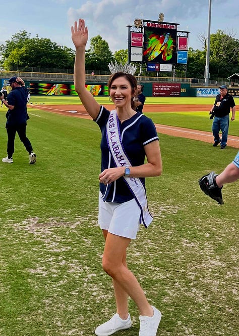 A woman wearing a blue dress and a jeweled crown stands smiling. She has a stethoscope around her neck, suggesting a medical profession, and holds a camouflage uniform jacket with a visible "U.S. AIR FORCE" patch over her shoulder. The background is a simple, light-colored gradient.