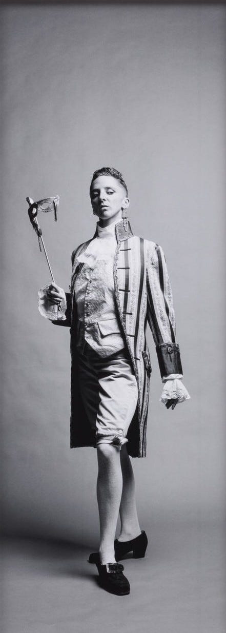 1) A black-and-white photo of a white woman with short dark hair, wearing large white feathered angel wings, a leather chest harness, and white shorts. The tone is dramatic and ethereal. She is standing to her side, with her left arm holding her stomach, and she is looking down and away from the camera. Her chest is bare, with the thin leather harness over it. 2) A woman in historical attire poses confidently in a black and white photo. They hold a feathered mask on a stick, wearing a striped coat and breeches. They have on traditional colonial shoes, and their white undershirt is long-sleeved with ruffled wrists. Her front left leg is out in front and turned to the side, while her chest is stuck out, and her left arm at her side, while the right arm holds the mask stick. She has short dark hair. 3) A woman in 18th-century attire poses elegantly in a black-and-white photograph. She wears a voluminous Victorian gown and a white powdered wig, holding a beaded mask in one hand and a sign in the other. The sign on her right reads: 'Una Troubridge, Alice Austen, Janet Flanner, Sappho, Radclyffe Hall'.