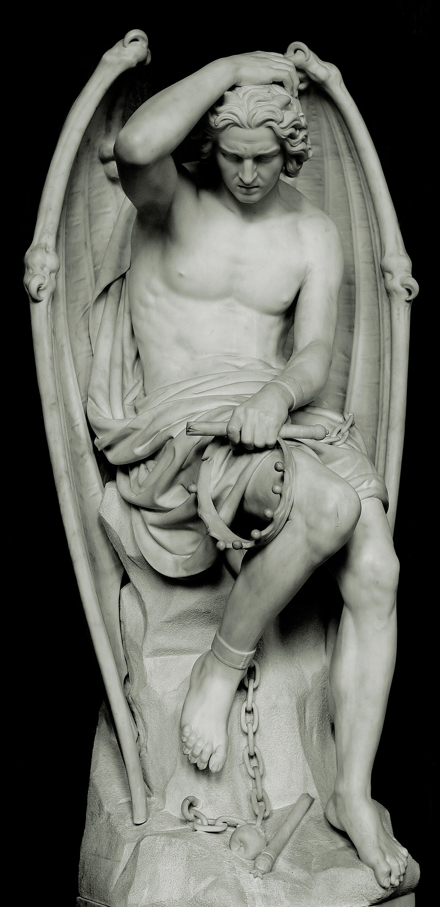 A white marble statue of a young winged man sitting on a rock, head bowed and chained, a broken crown at his feet, set at the back of a carved pulpit.