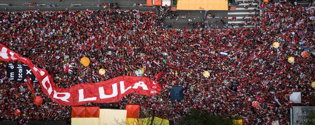 Multidão densa ocupa avenida em manifestação, com grande faixa vermelha escrita 'Lula'. Veículos e pedestres estão na rua, ladeados por edifícios altos.