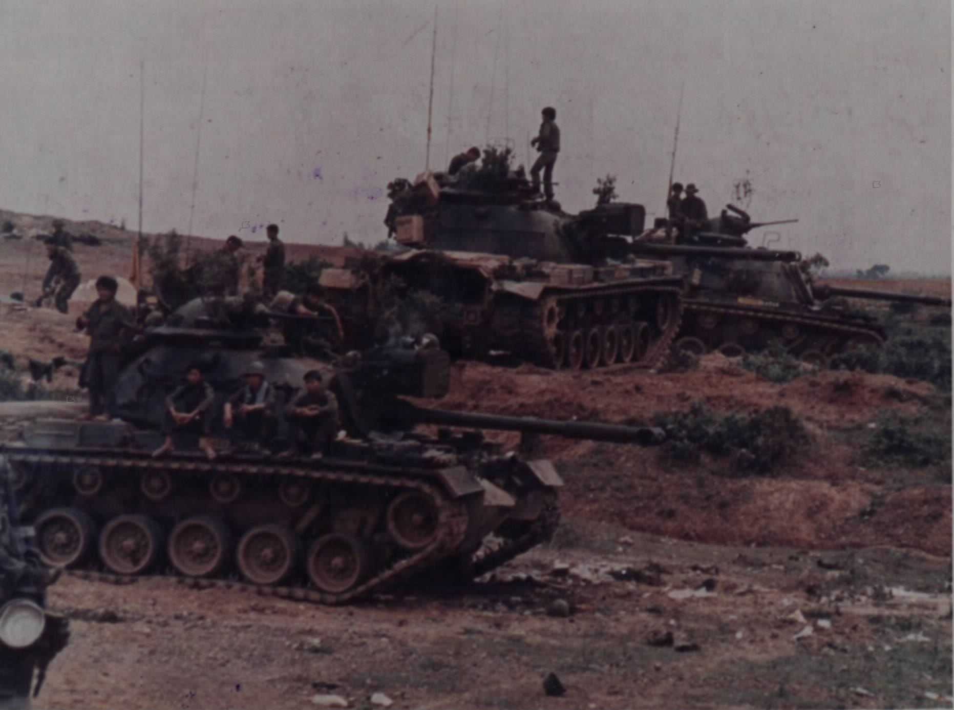 Two South Vietnamese M48 tanks with soldiers on top, parked on a riverbank under a grey sky. Two South Vietnamese M48 tanks with soldiers on top, parked on a riverbank under a grey sky.
