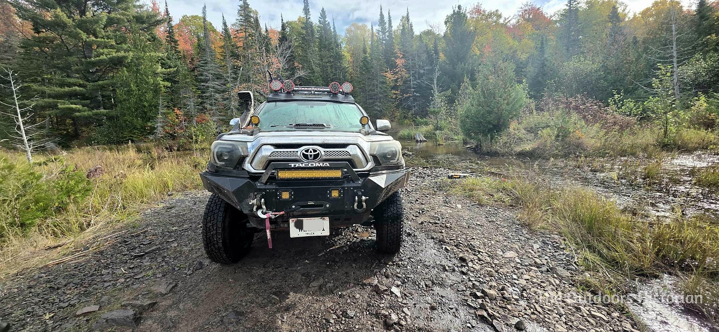 A head on view of a second generation black Toyota Tacoma with an added steel bumper, winch, snorkel, and multiple LED lights.