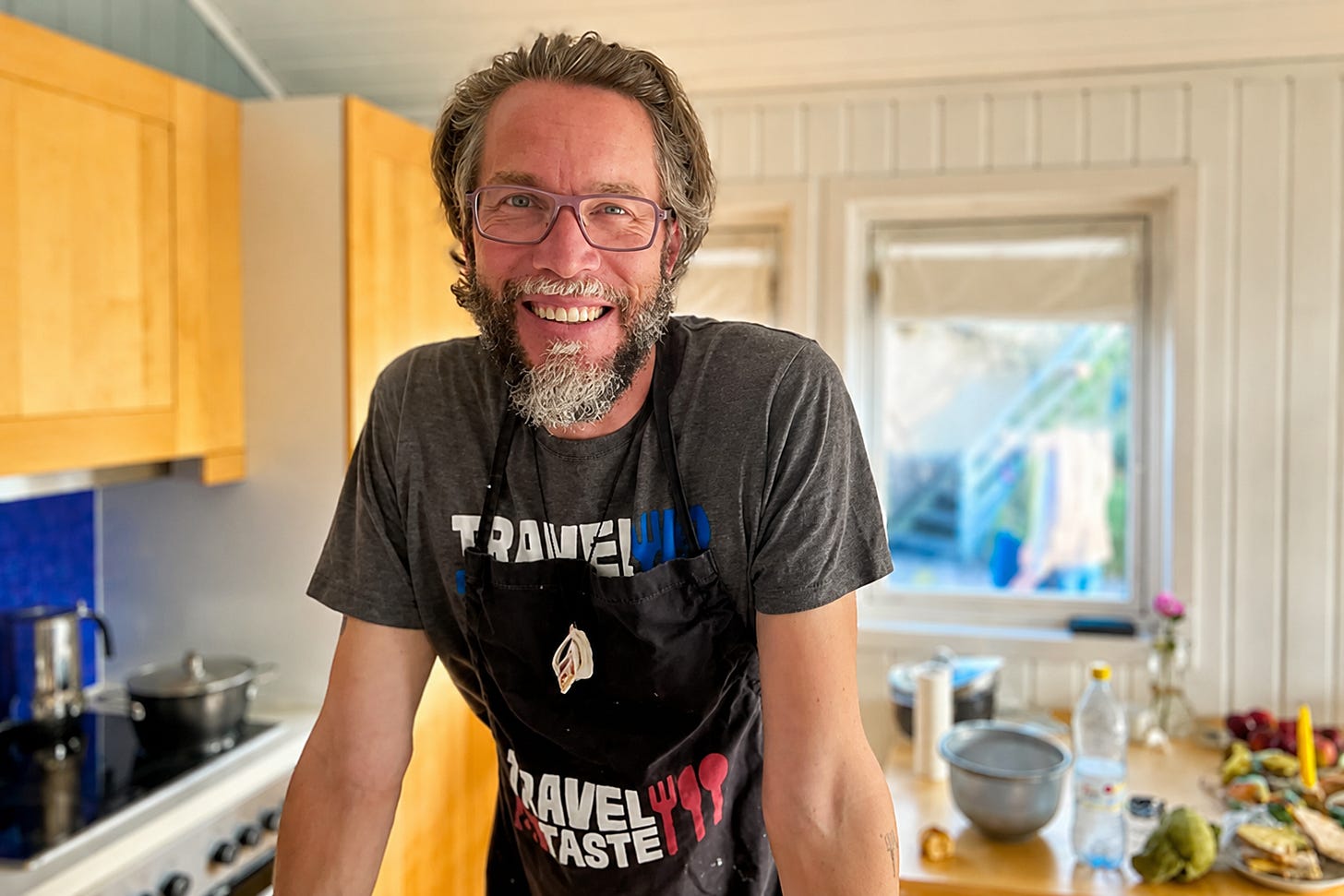 Patrick Hemminger mit Brille, Bart und schwarzer Schürze steht in einer hellen Küche und stützt sich auf die Arbeitsfläche. Im Hintergrund sind Herd, Kochtopf, Schüsseln, Gemüse und Fenster zu sehen, was die Szene wie eine entspannte Koch- oder Backsession wirken lässt.