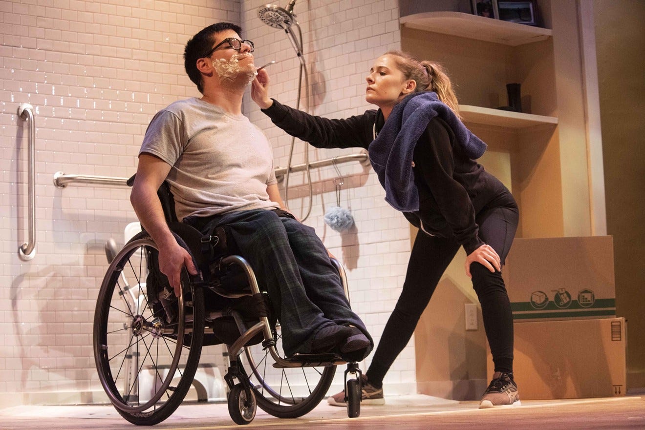 A white femme care worker shaves the face of a white masc wheelchair user against the backdrop of a shower.