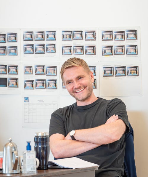 Adam Penford sitting at a wooden desk with a white wall with photos of a stage behind him