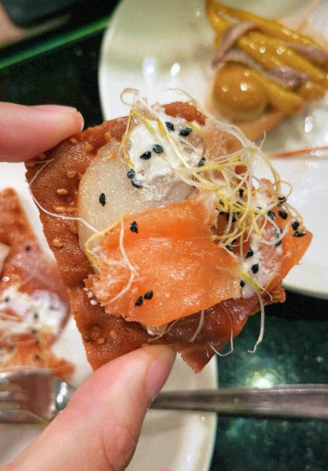 Crab stuffed squid, a spoon entering creamed uni in its shell, a hand holding a sesame crack with three different color pieces of smoked fish on top , a hand holding grilled foie on bread, a hand holding txistorra skewered onto a piece of bread, a fork entering a half eaten piece of cheesecake
