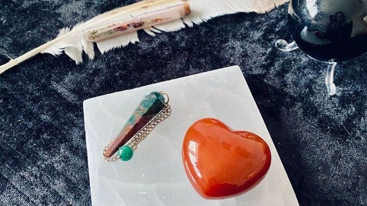 A witch’s altar with sacred and spellwork objects on a black velvet cloth covering (including a black tourmaline orb, barn owl feather, and goddess Athena spell jar. At the front is a quartz crystal charging plate, with a red/green bloodstone pendulum and a bright red/orange carnelian heart on top