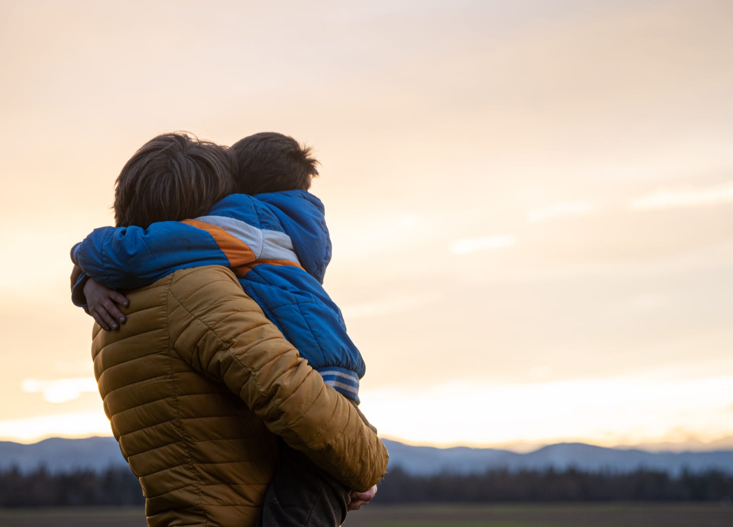 A father embracing his son as they stand together, facing a sunset over the mountains. A warm, reflective moment symbolizing connection, guidance, and the lasting impact of parenting.