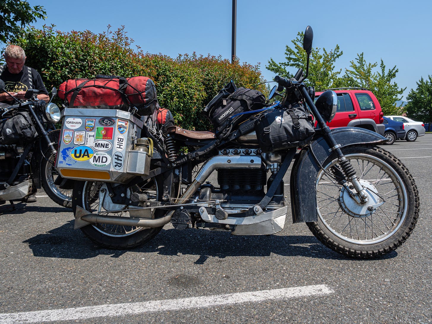 Photo of old Danish motorcycle outfited for a big adventure. The spoked wheels have knobby tires. There are aluminum panniers mounted on the back. The panniers are festooned with stickers from many places. On top of the aluminum boxes are red dry bags. There is a tank bag and a leather saddle.