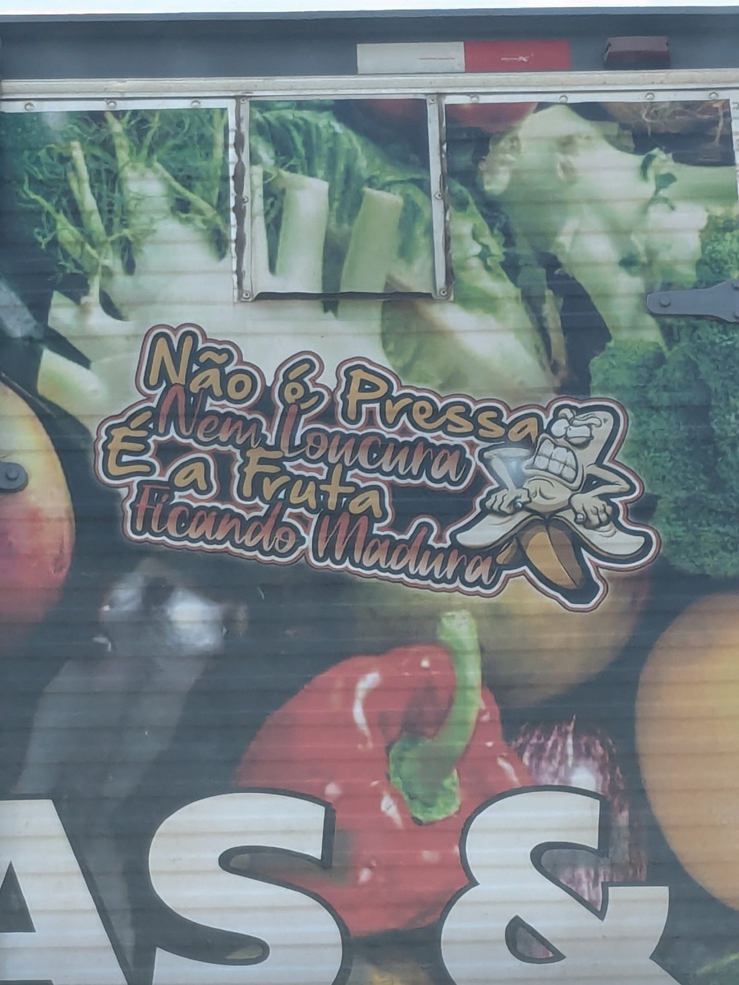 detalhe da parte traseira e um caminhão de frutas e verduras, que contém a seguinte frase: "não é pressa nem loucura, é a fruta ficando madura" e o desenho de uma banana saindo de dentro da casca com uma cara de desconforto, como se precisasse sair logo. no fundo, fotos de legumes diversos