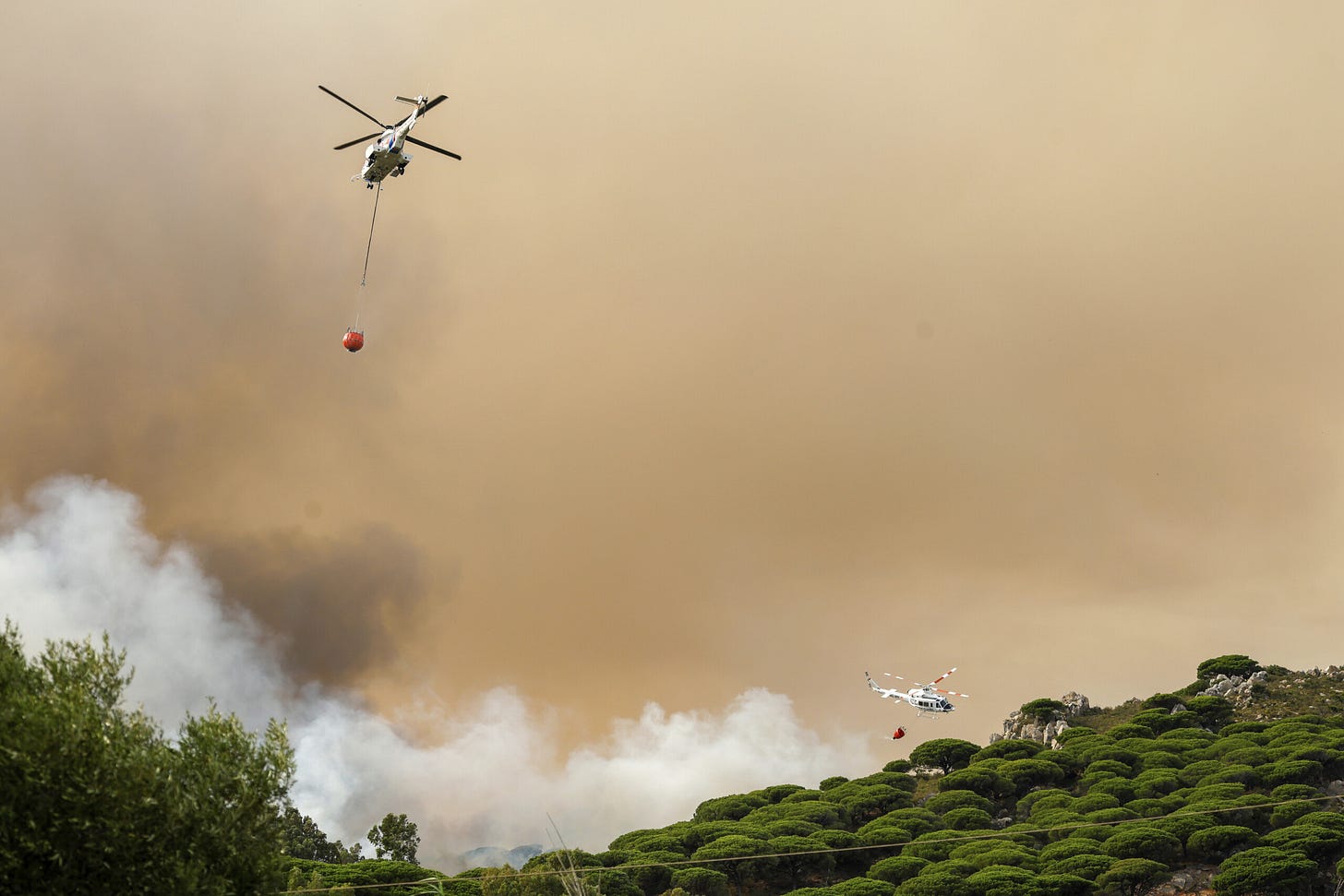 Two helicopters attempt to douse fires along a ridge. The sky is nicotene brown.