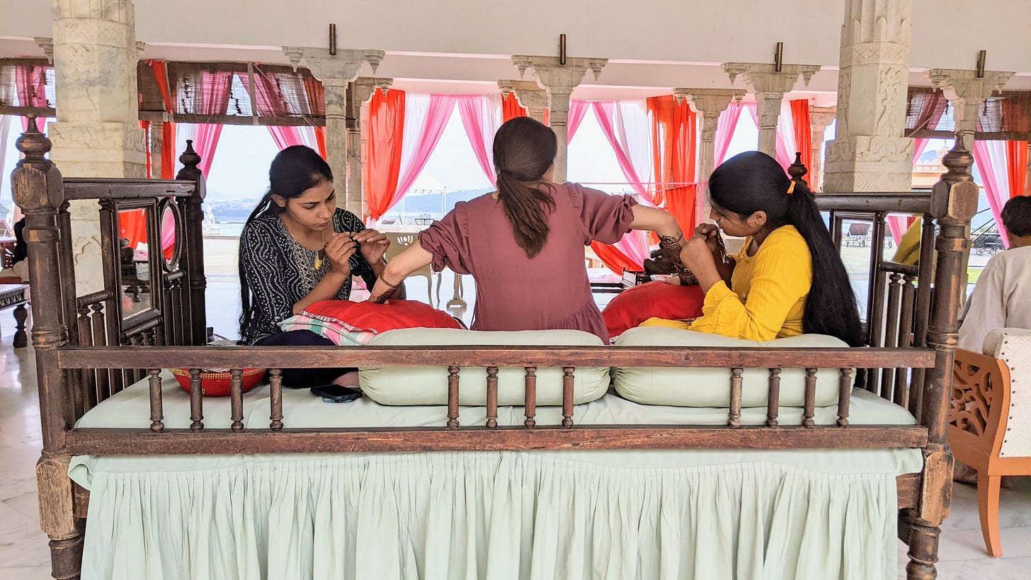 A brown-haired woman from behind, sitting between two other women who are applying henna to her arms