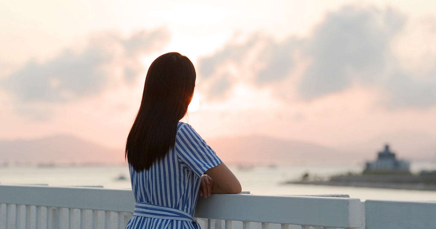 A woman with dark hair turns her back to the camera and stares pensively out to sea as she considers her true self.