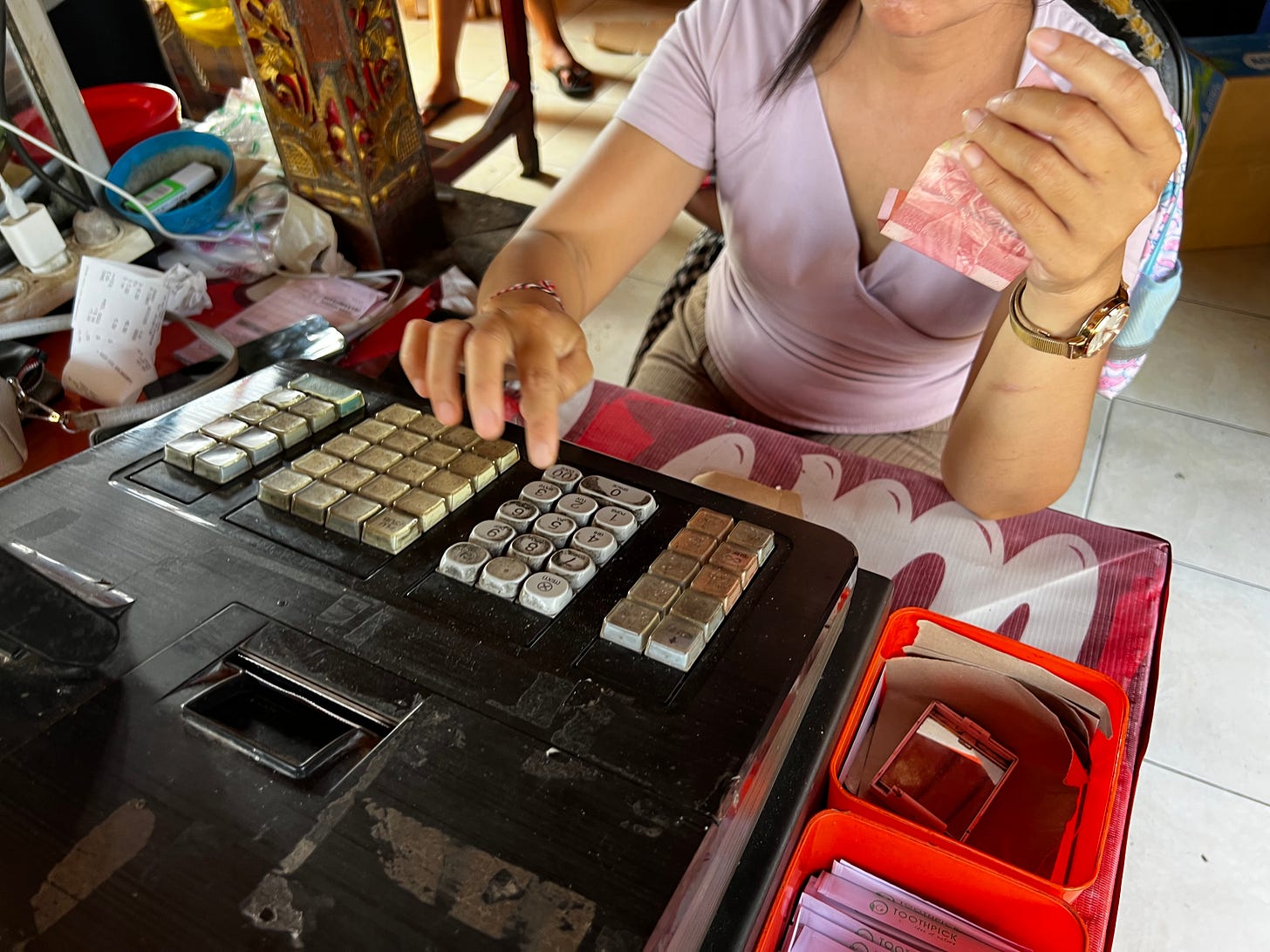 A hand making change at a worn cash drawer, holding a pink 100,000-rupiah note, a frayed red thread tied around the wrist.