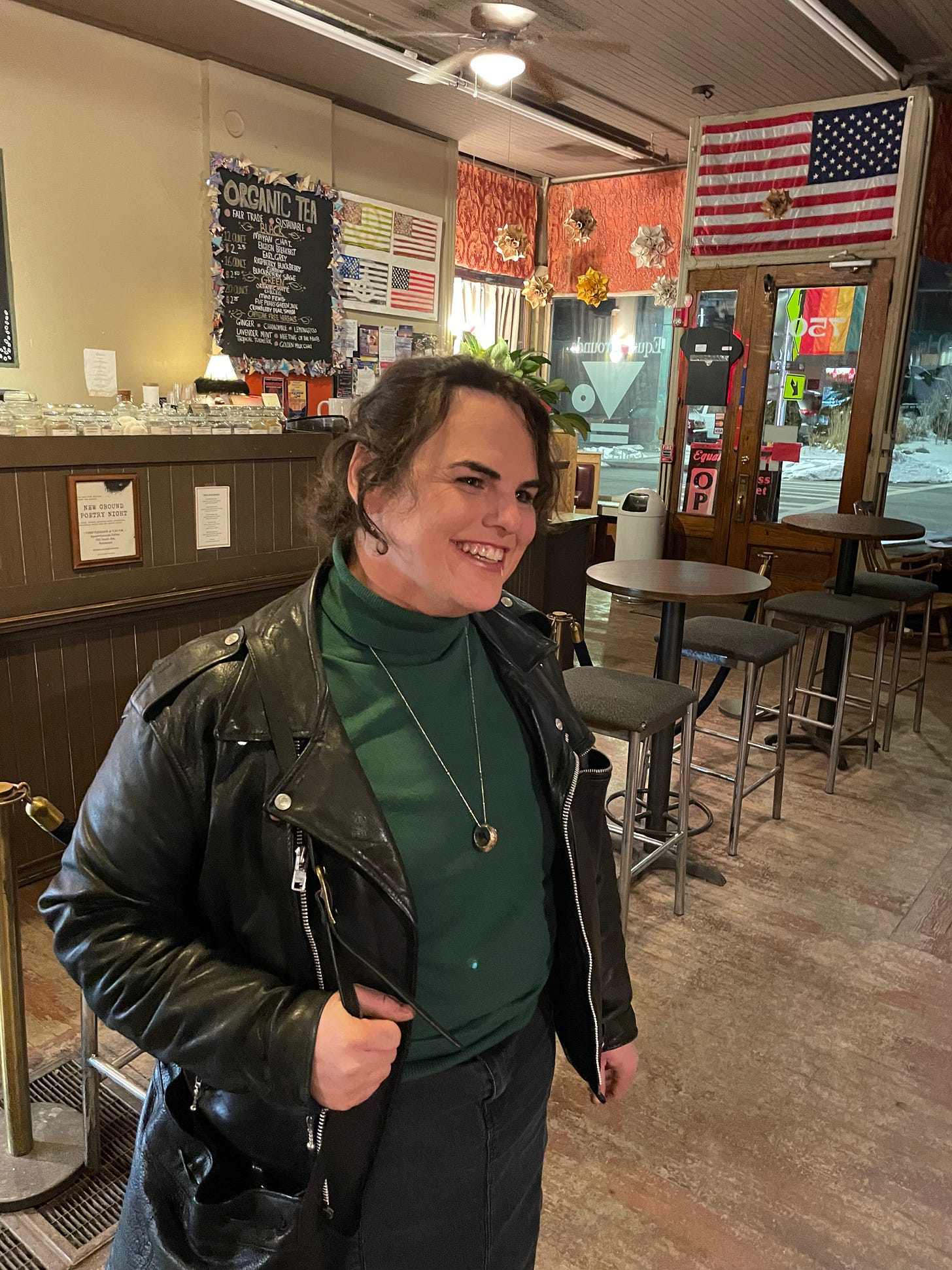 A smiling woman in a coffee shop; she's wearing the same green turtleneck in the previous photo, a leather jacket, and black denim skirt.  Various decorations, including an American flag and a Pride flag, can be seen in the background.