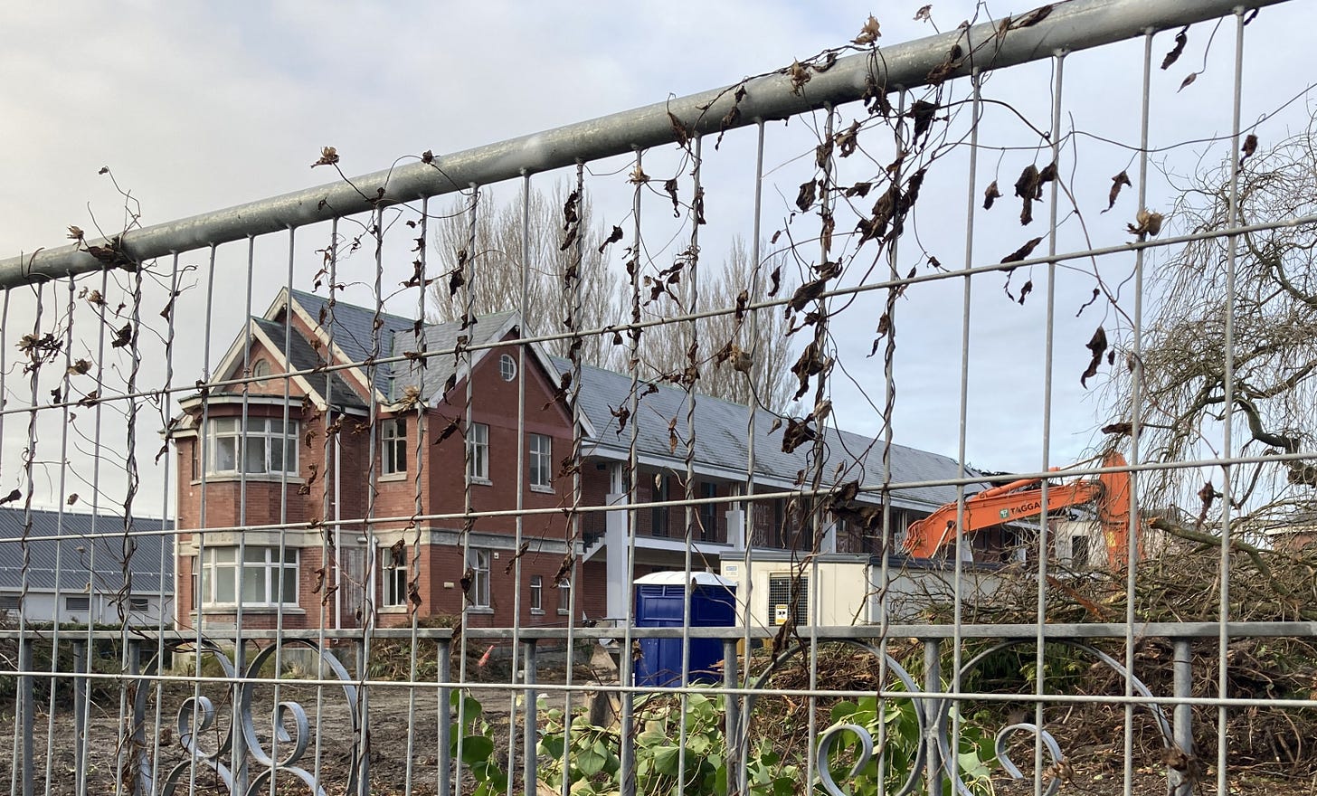 A dilapidated brick building, with a digger and portable toilet in the foreground, all behind a wire fence. A dilapidated brick building, with a digger and portable toilet in the foreground, all behind a wire fence.