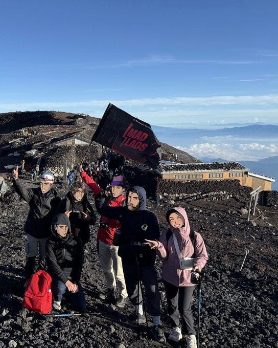 The image shows a group of people standing on the rocky summit of Mount Fuji, as indicated by the post text "Lads on Top (of Mt Fuji 🗻)" from Mad Monkey 🎒 (@MonkDoesnt). They are dressed in outdoor gear, including jackets and hiking poles, with one person holding a black flag with "Mad Lads" in red text. The background features a clear blue sky, distant mountains, and a building, likely a summit station, with clouds below, emphasizing the high altitude. The group appears joyful, posing for the photo, with a red backpack visible on the ground. No platform watermarks like Instagram, TikTok, or Xiaohongshu are present.