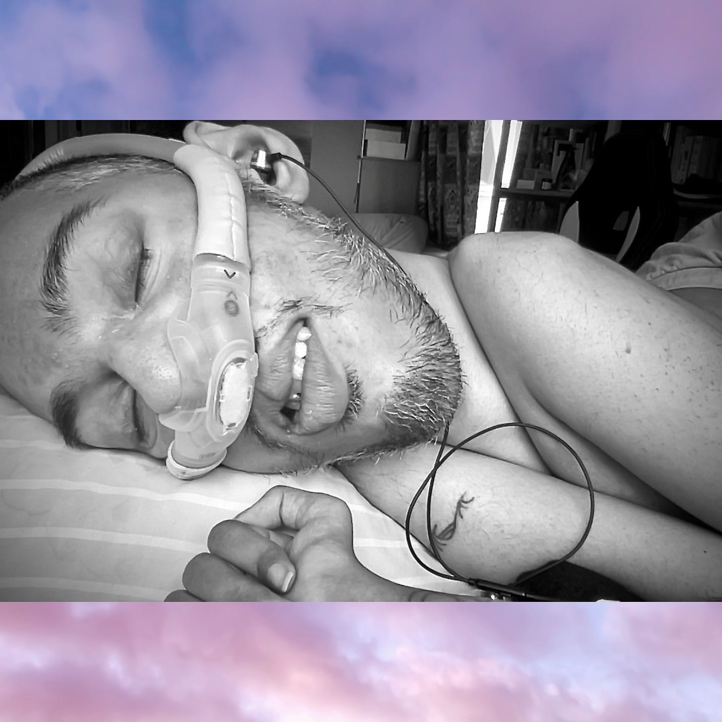 IMAGE DESCRIPTION: Bold, wild, fearless existence. On a background of purple clouds in the sunset, there is a black and white photo of Sulaiman (a wholeheartedly Disabled AF and British-Pakistani man with buzzcut black hair and facial stubble) lying in bed on his right side with his ventilator mask over his nose since he is done making his divine being palatable for an ableist world. His eyes are closed, and headphones are in his ear, listening to “Mending” by Frida Touray on loop as he takes a nap in the summer evening heat. He is shirtless with a wave tattoo on his right shoulder via Henry Hate, and Sulaiman wears pink shorts that look grey in the black and white photo. Crip/Wild/Sexy AF: The photo highlights Sulaiman’s Disabled AF and British-Pakistani body wasting by Muscular Dystrophy and twisting (and collapsing) by complex Scoliosis. Radical, restful, regenerative. DESCRIPTION ENDS.