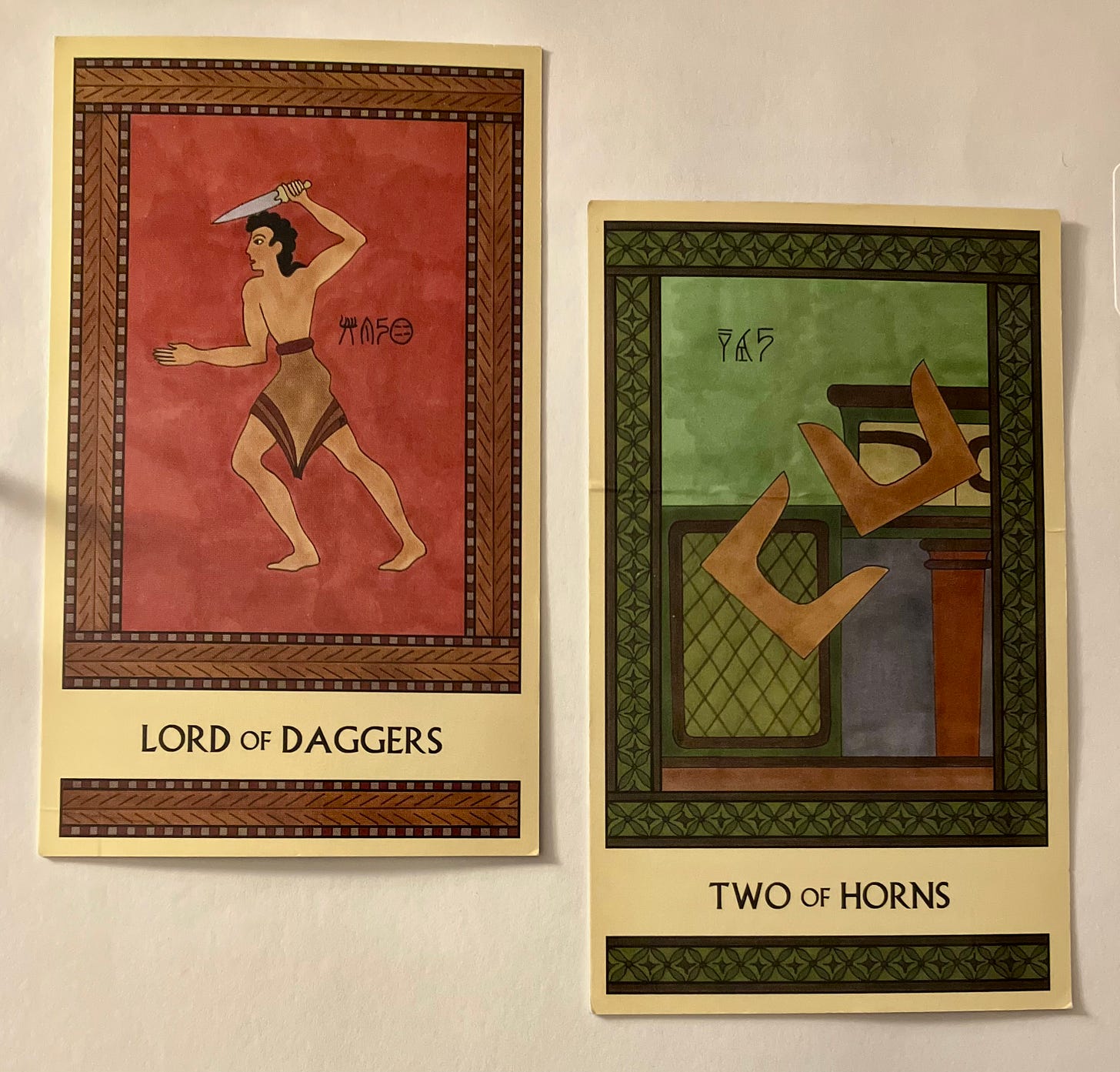 Two Minoan Tarot cards side by  side on a white surface. The Lord of Daggers, on the left, is painted in shades of deep red and brown. It shows a Minoan man wearing a short kilt, facing left in a dynamic pose, brandishing a dagger over his head. The Two of Horns, on the right, is painted in shades of deep green and brown. It shows two pairs of tan-colored sacred horns in midair as they tumble off a shrine. 