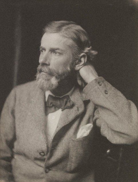 Black-and-white portrait of Edward Carpenter seated in three-quarter profile, resting one hand behind his neck. He wears a suit jacket with a bow tie and looks thoughtfully off to the side, his beard neatly trimmed and hair swept back.