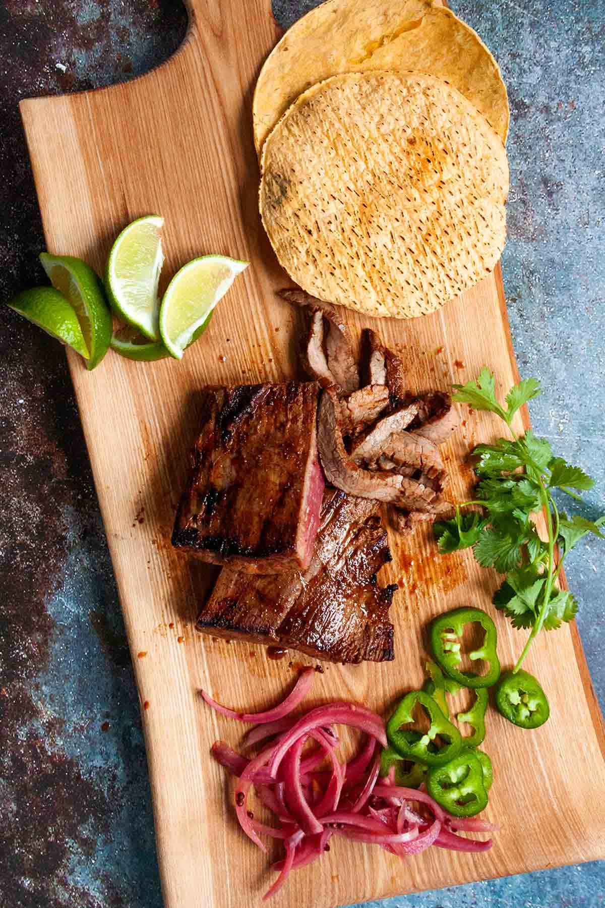 A cutting board with slices of Mexican skirt steak, pickled red onions, jalapeño, and three corn tortillas.