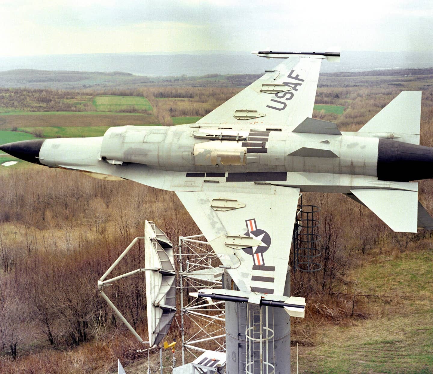 An F-16 Fighting Falcon aircraft is mounted upside-down onto a 30-foot pedestal at the Rome Air Development Center's Newport Site, May 1983, for evaluation of the ASPJ and ALR-69 antenna systems. From the positioner, the aircraft can be tested as if it were in flight without the high coast of flight testing. <em>National Archives</em>